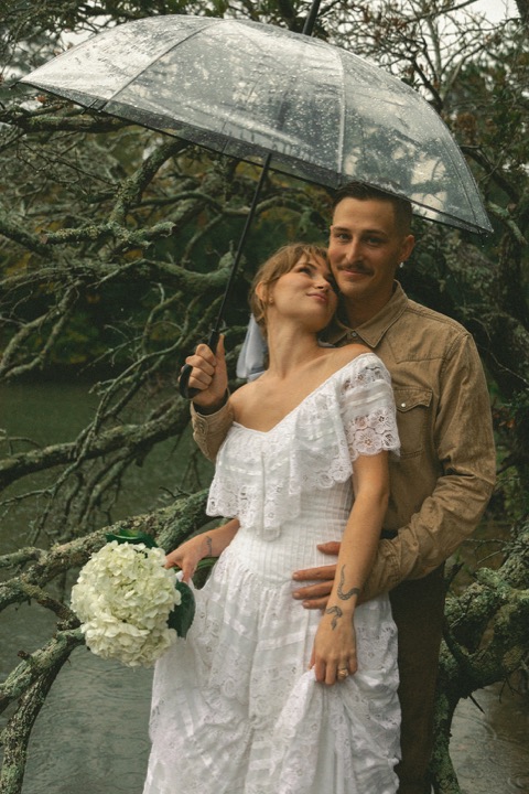 The couple smiling beneath a clear umbrella in the gardens
