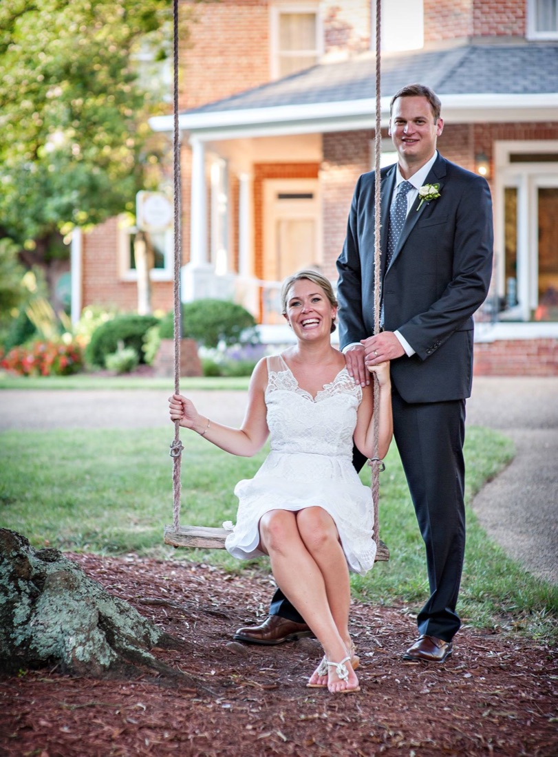 A couple posing on the garden swing beneath a tree, with the Manor's porches in the background