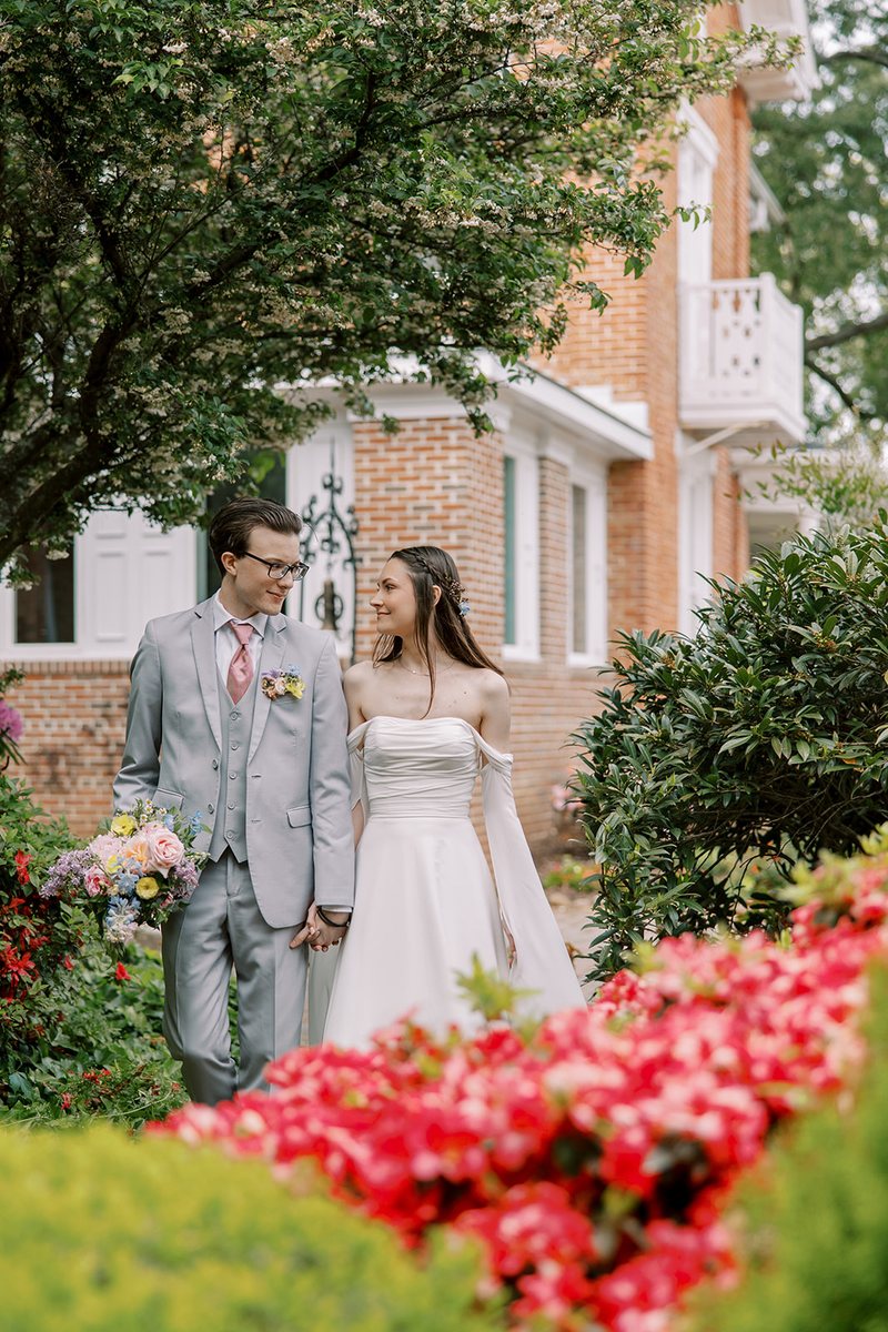 A couple holding hands among flowering gardens beside the historic brick Manor